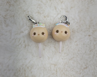 May include: Two small, brown, round cake pops with white sticks and white frosting topped with colorful sprinkles. The cake pops have cute, hand-painted faces with brown eyes and pink cheeks.