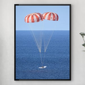 May include: A framed print of a spacecraft descending into the ocean. Two red and white striped parachutes are deployed above the water. The image is set against a blue sky and ocean backdrop.