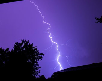 Könnte beinhalten: Ein heller, weißer Blitz streift über einen dunkelvioletten Himmel, vor dem sich Bäume und ein Dach abzeichnen.
