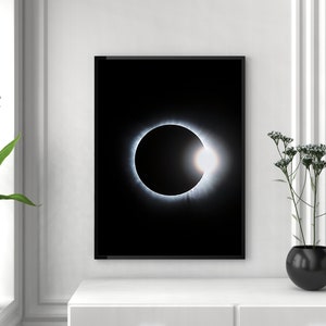 May include: A black and white photograph of a solar eclipse. The sun is completely covered by the moon, with only a thin ring of light visible around the edges. The photo is framed in a black frame and hanging on a white wall.