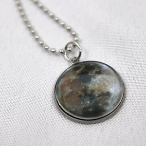 May include: A round pendant necklace featuring a detailed image of the moon. The pendant has a silver-colored metal frame and hangs from a ball chain necklace. The moon image displays shades of gray, brown, and blue.