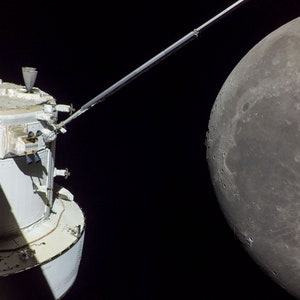 May include: A close-up view of a spacecraft in orbit with the moon in the background. The spacecraft is white with a long, thin arm extending out to the right. The moon is a large, gray sphere with a dark, cratered surface.