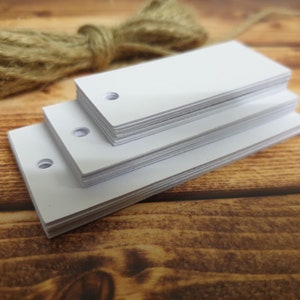 May include: A stack of white rectangular gift tags with a hole punched in the top. The tags are made of sturdy cardstock and are perfect for adding a personal touch to gifts.