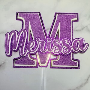 May include: A purple glitter cake topper featuring the letter "M" and the name "Merissa" in a cursive font. The topper has white and gold accents and is attached to a clear plastic stick.