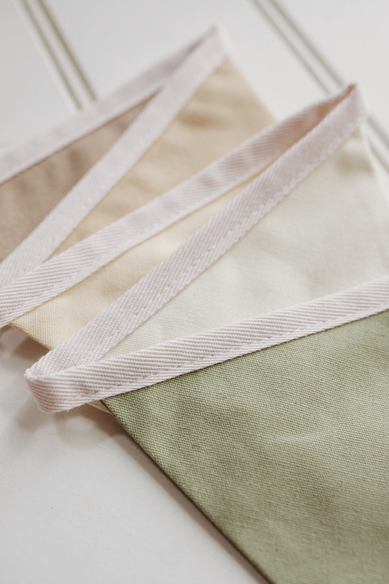 May include: A set of fabric pennants with a white, beige, and green color scheme. The pennants have a white trim around the edges.