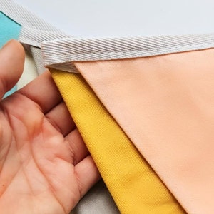 K&ouml;nnte beinhalten: Ein farbenfrohes Stoffbanner mit dreieckigen Flaggen in den Farben Pfirsich, Gelb, Grau und Hellblau. Die Flaggen sind mit wei&szlig;em Stoffbesatz zusammengen&auml;ht.