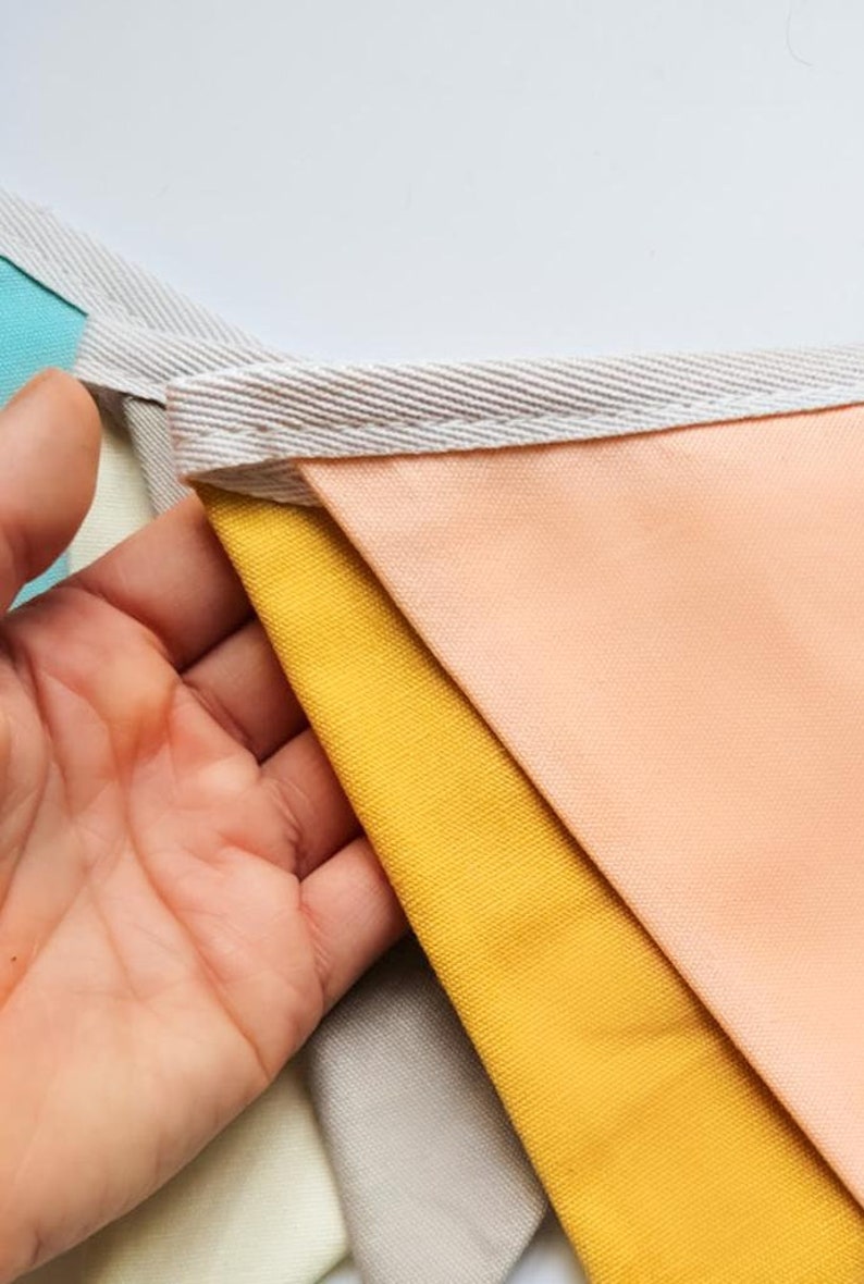 K&ouml;nnte beinhalten: Ein farbenfrohes Stoffbanner mit dreieckigen Flaggen in den Farben Pfirsich, Gelb, Grau und Hellblau. Die Flaggen sind mit wei&szlig;em Stoffbesatz zusammengen&auml;ht.