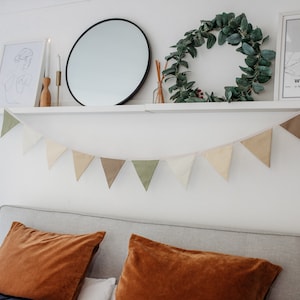 May include: A white shelf with a round mirror, a wreath of green leaves, a framed print with the text "Whitstable, United Kingdom", and two candlesticks. A string of triangular fabric bunting hangs below the shelf in shades of green, brown, and beige. The bunting is hanging above a gray headboard with two orange velvet pillows.