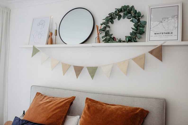 May include: A white shelf with a round mirror, a wreath of green leaves, a framed print with the text "Whitstable, United Kingdom", and two candlesticks. A string of triangular fabric bunting hangs below the shelf in shades of green, brown, and beige. The bunting is hanging above a gray headboard with two orange velvet pillows.