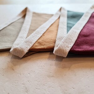 May include: Close-up of a colorful bunting banner with triangular flags in shades of beige, gold, teal, and burgundy. Each flag is trimmed with a cream-colored woven border. The banner is displayed on a light-colored surface.
