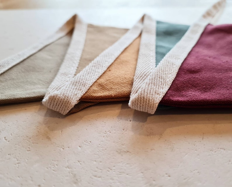 May include: Close-up of a colorful bunting banner with triangular flags in shades of beige, gold, teal, and burgundy. Each flag is trimmed with a cream-colored woven border. The banner is displayed on a light-colored surface.