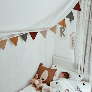 May include: A child's bedroom scene with a white bed, bedding with tree and house motifs, and a brown bear-shaped pillow. A garland of triangular flags in various earth tones hangs above the bed. A small child sleeps in the bed.