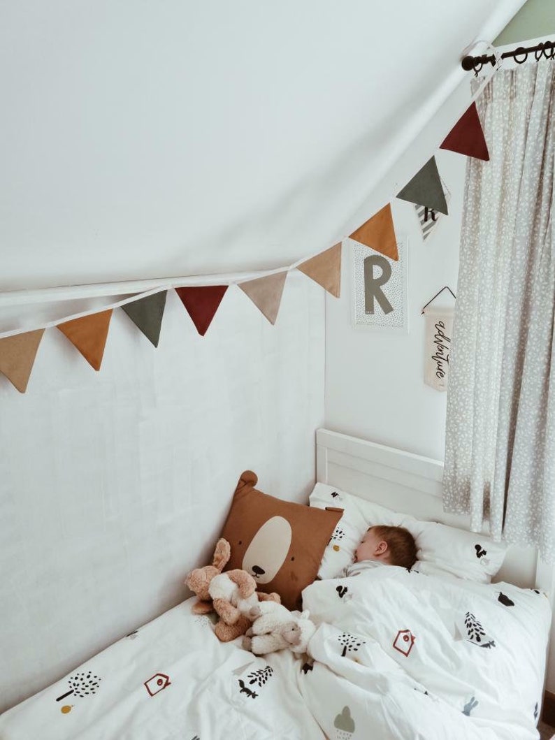 May include: A child's bedroom scene with a white bed, bedding with tree and house motifs, and a brown bear-shaped pillow. A garland of triangular flags in various earth tones hangs above the bed. A small child sleeps in the bed.