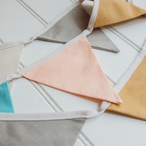 K&ouml;nnte beinhalten: Eine Girlande mit Flaggen in verschiedenen Farben, darunter Beige, Hellblau, Pfirsich und Senfgelb. Die Flaggen sind mit wei&szlig;em Stoff zusammengen&auml;ht.