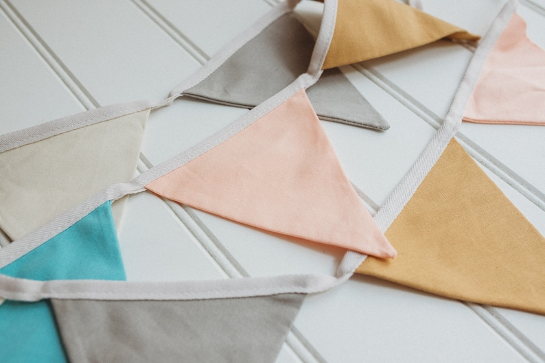 K&ouml;nnte beinhalten: Eine Girlande mit Flaggen in verschiedenen Farben, darunter Beige, Hellblau, Pfirsich und Senfgelb. Die Flaggen sind mit wei&szlig;em Stoff zusammengen&auml;ht.