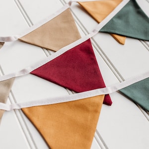 May include: A colorful bunting banner with triangular flags in shades of tan, burgundy, mustard yellow, and green. Each flag is trimmed with white fabric and strung together on a white ribbon. The banner is displayed on a white, slatted surface.