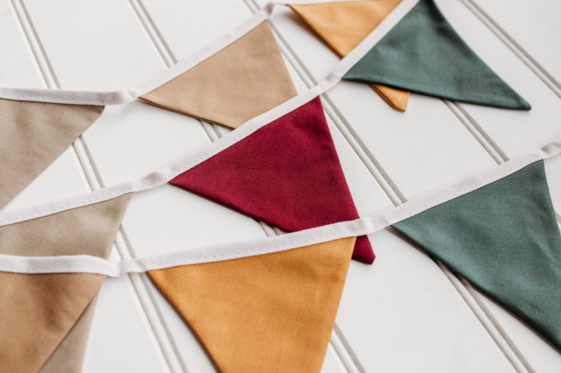May include: A colorful bunting banner with triangular flags in shades of tan, burgundy, mustard yellow, and green. Each flag is trimmed with white fabric and strung together on a white ribbon. The banner is displayed on a white, slatted surface.