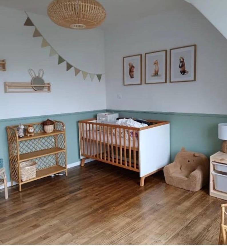May include: A white and wood crib with a white and pink mattress, a wicker shelf with toys, a brown stuffed animal chair, and three framed animal prints on the wall. The room is decorated in a woodland theme with a green and white color scheme.