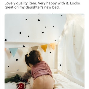 K&ouml;nnte beinhalten: Ein kleines Kind spielt in einem Bett mit einem wei&szlig;en Baldachin. Das Bett hat eine wei&szlig;e Wand mit schwarzen Punkten und eine Girlande mit bunten Flaggen, die &uuml;ber dem Bett h&auml;ngt.