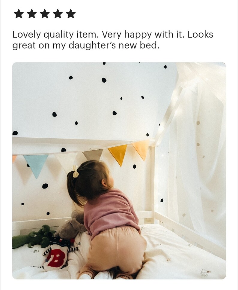 K&ouml;nnte beinhalten: Ein kleines Kind spielt in einem Bett mit einem wei&szlig;en Baldachin. Das Bett hat eine wei&szlig;e Wand mit schwarzen Punkten und eine Girlande mit bunten Flaggen, die &uuml;ber dem Bett h&auml;ngt.