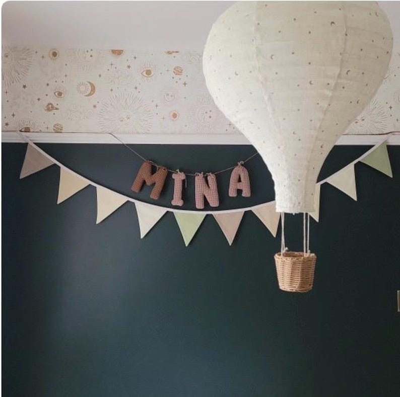 May include: A white hot air balloon with brown crocheted letters spelling "MINA" hanging from a string of flags. The balloon is hanging from the ceiling against a dark green wall.