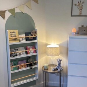 May include: White bookshelf with four shelves, filled with children's books and stuffed animals. The bookshelf is mounted on a wall with a green and white arch painted behind it. The bookshelf has a wooden rail on each shelf to keep the books in place.