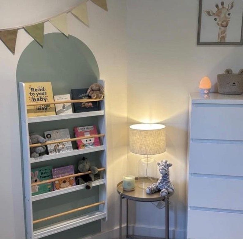 May include: White bookshelf with four shelves, filled with children's books and stuffed animals. The bookshelf is mounted on a wall with a green and white arch painted behind it. The bookshelf has a wooden rail on each shelf to keep the books in place.