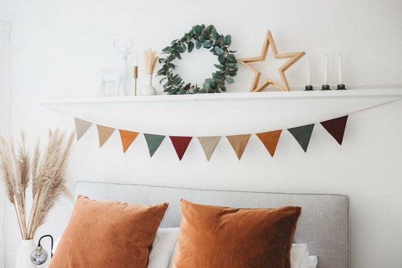 May include: A cozy bedroom scene featuring a neutral color palette. A garland of triangular flags in various earth tones hangs above a bed with orange pillows. A shelf displays a wreath, wooden star, candles, and decorative items.