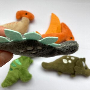 May include: Five plush dinosaur toys, including a gray stegosaurus with green spines, a green dinosaur, a brown dinosaur with an orange head, an orange pterodactyl, and a green triceratops.