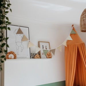 May include: A nursery room with a white wall, a brown woven pendant light, a white shelf with a framed print of a child, a wooden name sign, and a peach-colored canopy bed with a triangle garland.