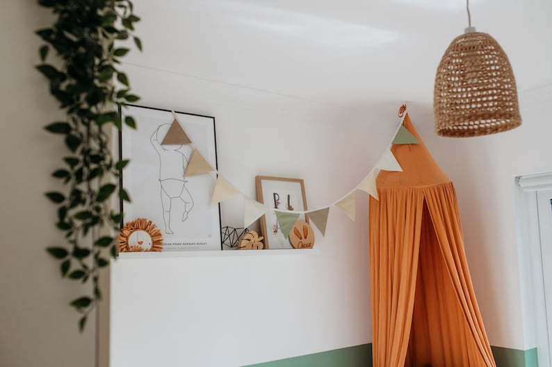 May include: A nursery room with a white wall, a brown woven pendant light, a white shelf with a framed print of a child, a wooden name sign, and a peach-colored canopy bed with a triangle garland.