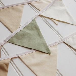 May include: A bunting with flags in shades of beige, green, and white. The flags are sewn together with a white ribbon.