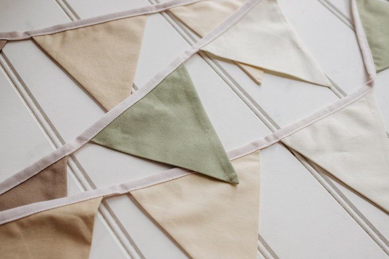 May include: A bunting with flags in shades of beige, green, and white. The flags are sewn together with a white ribbon.