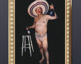 May include: A man wearing a large, wide-brimmed straw sombrero with a red and white band, holding a bottle of liquor in one hand and a glass in the other. He is wearing a pair of plaid swim trunks and has a mustache and beard.