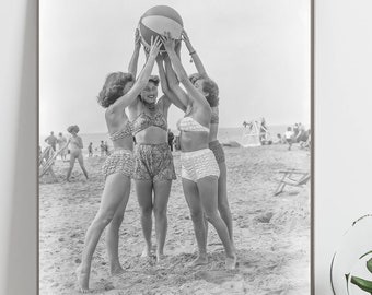 May include: Black and white photograph of four women on a beach, tossing a beach ball in the air. The women are wearing bathing suits and are smiling. The beach is crowded with people.