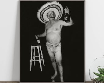 May include: Black and white photograph of a man wearing a sombrero and holding a bottle of whiskey. He is standing in front of a bar stool and wearing a pair of checkered swim trunks.
