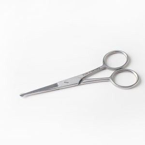 May include: Silver-toned grooming scissors with rounded tips. The scissors have a small hole in the center of the handle and the words "Made in Germany" are engraved on the handle.