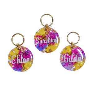 Puede incluir: Tres llaveros redondos con herrajes dorados. Cada llavero presenta una base de resina transparente llena de flores secas amarillas, moradas y rosas. Los llaveros tienen los nombres "Chloe", "Sunshine" y "Gilda" en escritura blanca.