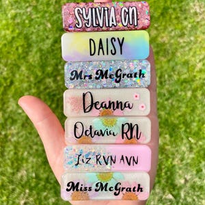 Puede incluir: Una pila de siete placas de identificación rectangulares de resina con varios diseños, incluyendo purpurina rosa, degradados pastel, copos holográficos y patrones florales. Cada placa muestra un nombre o título en letras negras: Sylvia RN, Daisy, Mrs. McGrath, Deanna, Octavia RN, Liz RVN AVN y Miss McGrath.