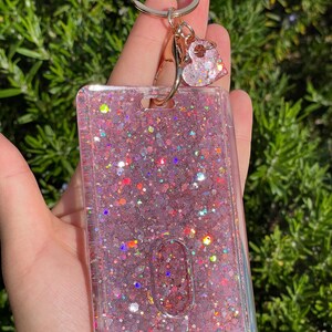 May include: A rectangular clear plastic ID badge holder filled with pink glitter and iridescent sequins. The holder features a gold-toned metal keyring attachment with a small, matching heart-shaped charm dangling from the top clasp.