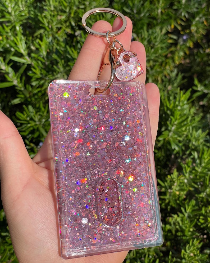 May include: A rectangular clear plastic ID badge holder filled with pink glitter and iridescent sequins. The holder features a gold-toned metal keyring attachment with a small, matching heart-shaped charm dangling from the top clasp.