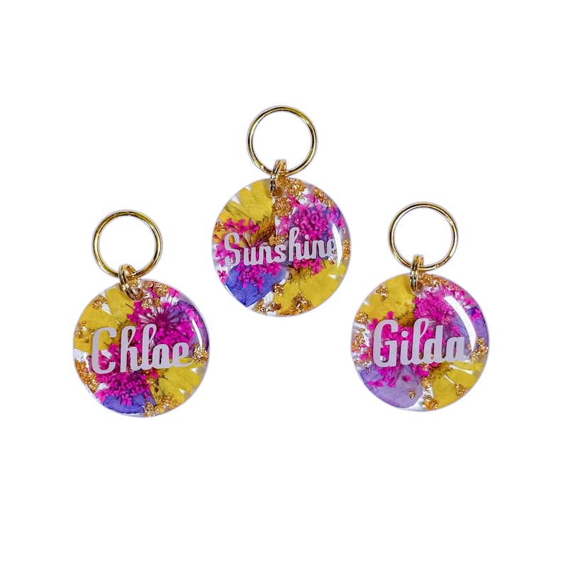 Puede incluir: Tres llaveros redondos con herrajes dorados. Cada llavero presenta una base de resina transparente llena de flores secas amarillas, moradas y rosas. Los llaveros tienen los nombres "Chloe", "Sunshine" y "Gilda" en escritura blanca.