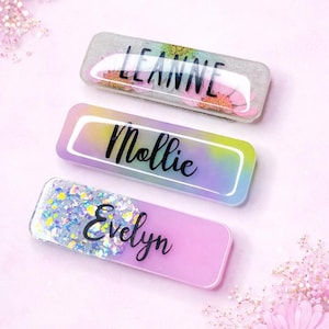 May include: Three rectangular name badges with rounded edges. The top badge is clear with the name "LEANNE" in black script, with floral accents. The middle badge has the name "Mollie" in black script. The bottom badge is pink with the name "Evelyn" in black script.