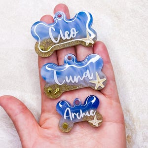 May include: Three blue and white bone-shaped dog tags with gold glitter and seashells. The tags are personalised with the names Cleo, Luna, and Archie.