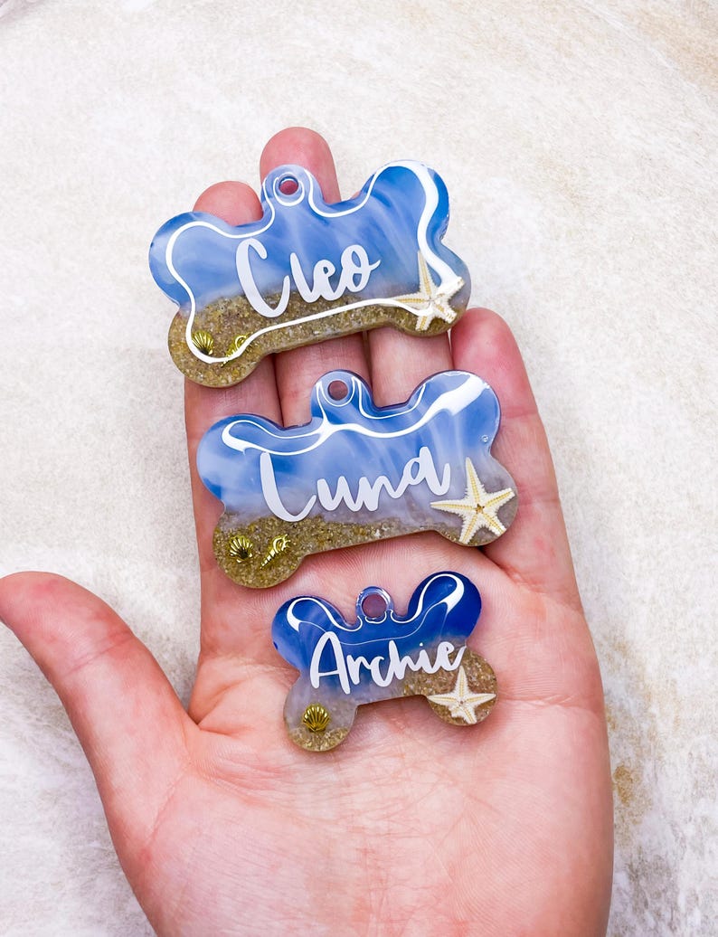 May include: Three blue and white bone-shaped dog tags with gold glitter and seashells. The tags are personalised with the names Cleo, Luna, and Archie.