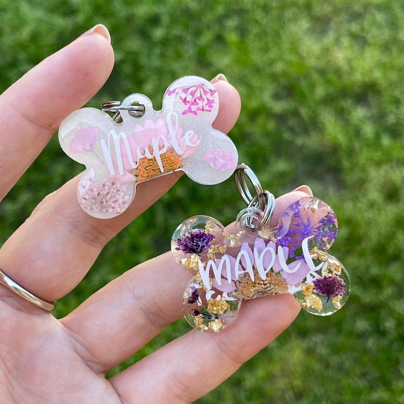 Puede incluir: Dos etiquetas para mascotas en forma de hueso con el nombre "Maple" en escritura blanca. Las etiquetas son de resina transparente con flores secas y copos de oro. Una etiqueta tiene flores rosas y naranjas, la otra moradas y doradas.