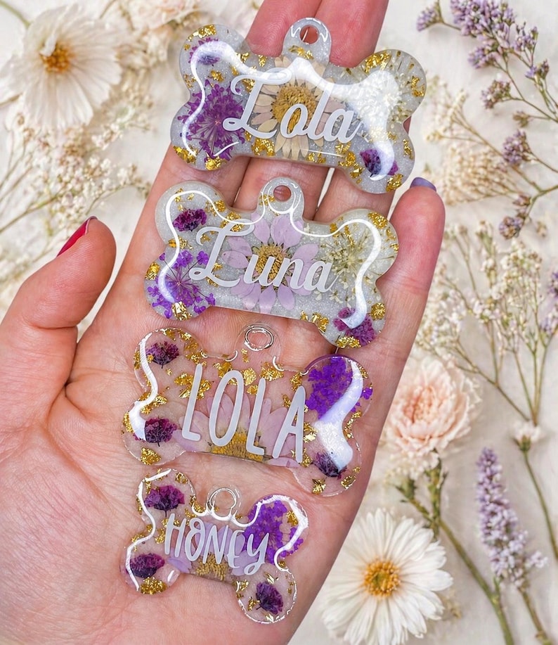 Puede incluir: Cuatro etiquetas para perros de resina transparente en forma de hueso, cada una con un nombre diferente en escritura blanca: Lola, Luna, Lola y Honey. Las etiquetas est&aacute;n llenas de flores secas y copos de oro. El fondo incluye flores blancas y moradas.