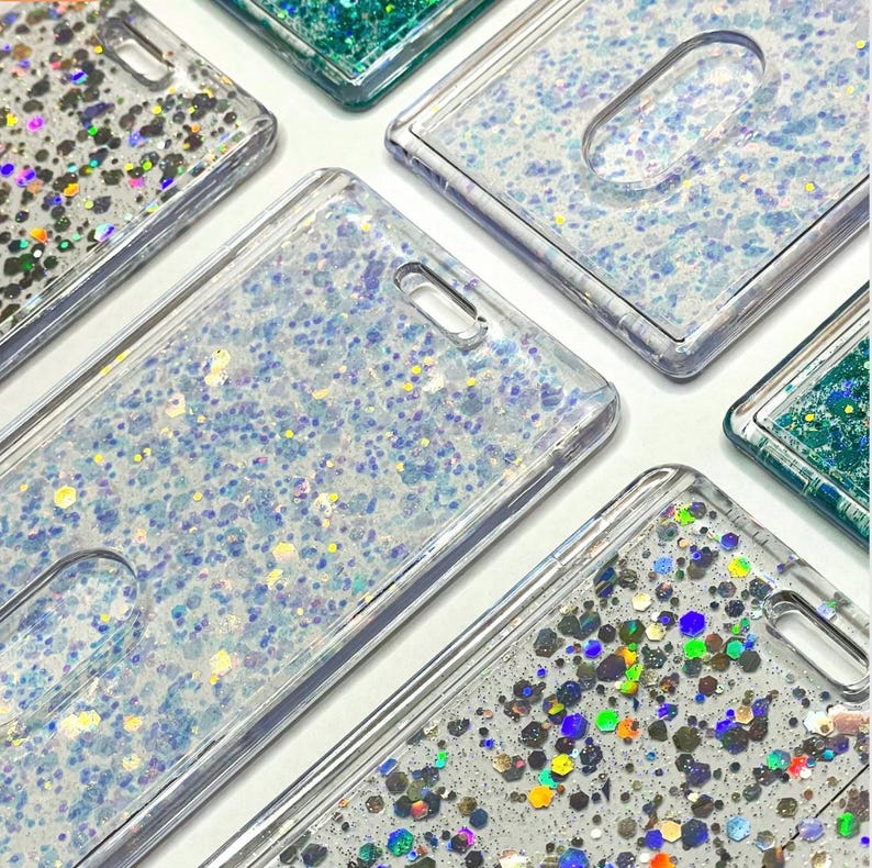 May include: A collection of rectangular clear plastic badge holders featuring liquid-filled interiors with floating glitter. Some holders contain fine iridescent blue and white glitter, while others feature larger, multi-colored hexagonal sequins.
