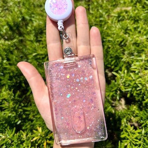 May include: A rectangular clear plastic ID badge holder filled with pink liquid and iridescent glitter, attached to a white retractable badge reel featuring a matching pink glitter center. The holder has a vertical slot for a card and a metal clip.
