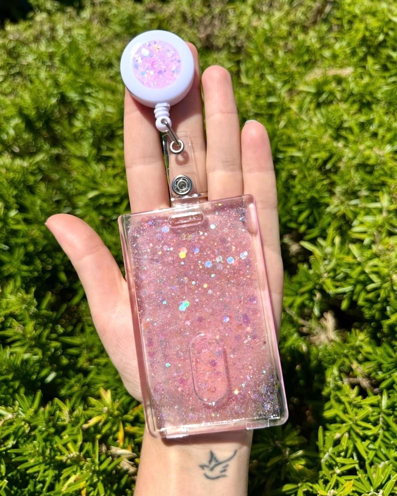 May include: A rectangular clear plastic ID badge holder filled with pink liquid and iridescent glitter, attached to a white retractable badge reel featuring a matching pink glitter center. The holder has a vertical slot for a card and a metal clip.